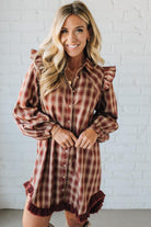 Woman wearing a plaid dress with ruffled sleeves and hem against a white brick wall.