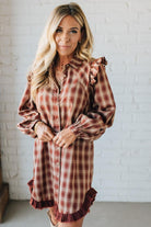 Woman wearing a plaid dress with ruffled sleeves and hem against a white brick wall.