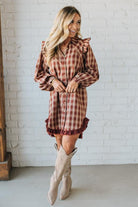 Woman wearing a plaid dress with ruffled details and beige knee-high boots against a white brick wall.