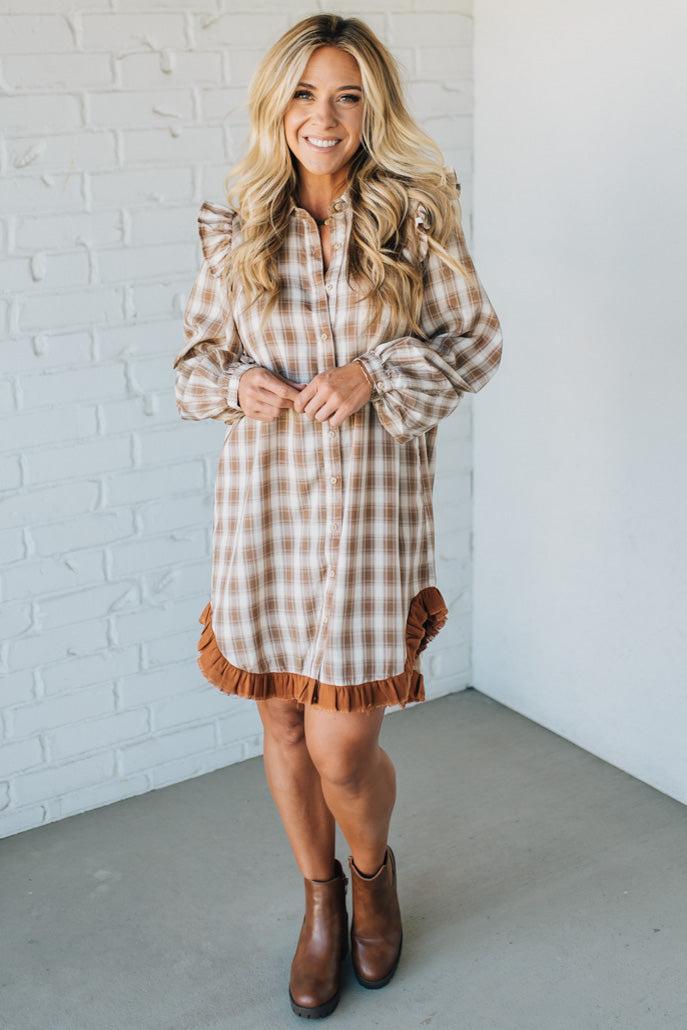 Woman wearing a plaid dress with brown trim against a white brick wall.
