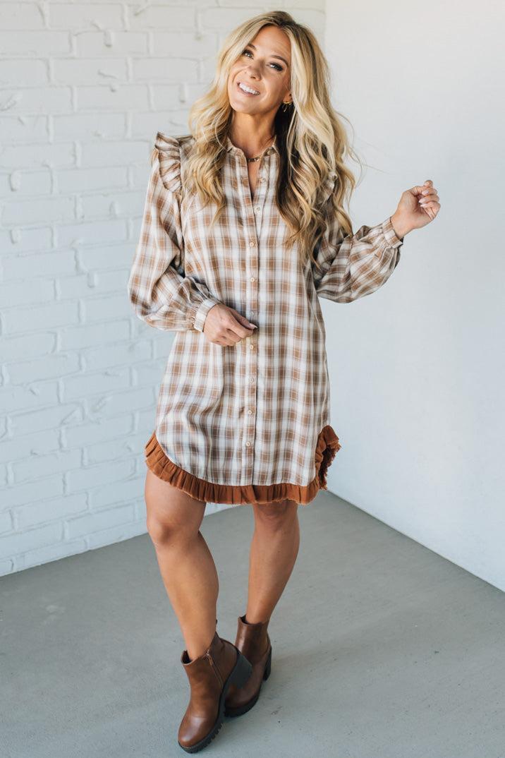 Woman wearing a plaid dress with ruffled details against a white brick wall.