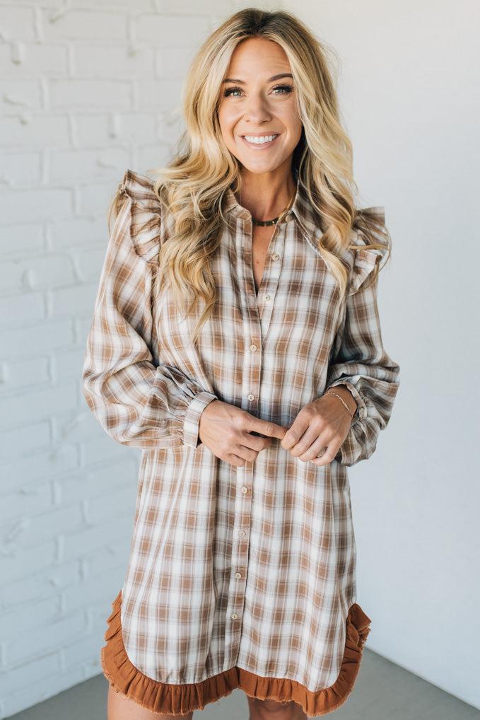 Woman wearing a plaid dress with ruffled details against a white brick wall.