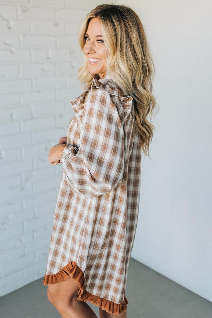 Woman wearing a plaid dress with ruffled details against a white brick wall.