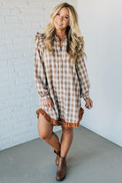 Woman wearing a plaid dress with ruffled hem and boots against a white brick wall.