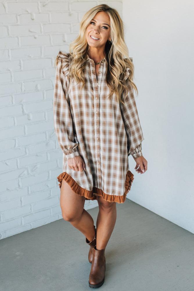 Woman wearing a plaid dress with ruffled hem and boots against a white brick wall.