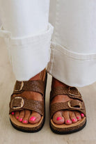 Brown sandals with gold buckles worn with white pants on a light wooden floor.