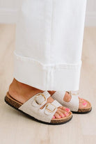 White sandals with gold buckles worn by a person with pink nail polish on a light wooden floor.