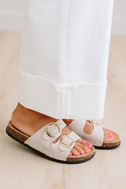 White sandals with gold buckles worn by a person with pink nail polish on a light wooden floor.
