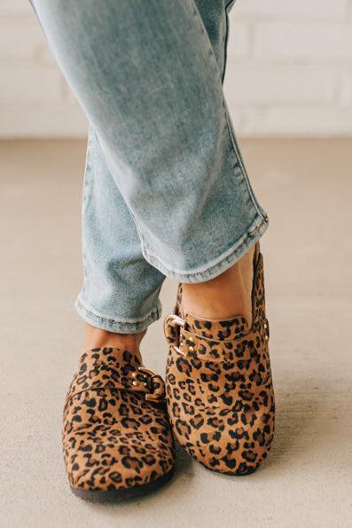 Woman wearing leopard print faux leather slip on clogs with single gold buckle on the side.