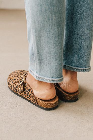 Woman wearing leopard print faux leather slip on clogs with single gold buckle on the side.