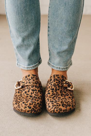 Woman wearing leopard print faux leather slip on clogs with single gold buckle on the side.
