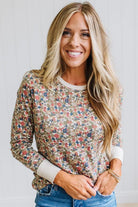 A smiling blonde woman wearing a long-sleeve crewneck sweatshirt with a dense multicolor floral pattern on a beige background, paired with blue jeans.