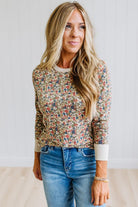 A woman with long blonde hair wearing a long-sleeve crewneck top with a dense multicolor floral print on a beige base, paired with blue jeans, standing against a white paneled wall.