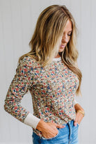 A woman wearing a beige long-sleeve crewneck sweatshirt with a dense multicolor floral print, paired with blue jeans, standing against a white paneled wall.