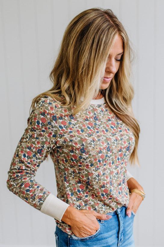 A woman wearing a beige long-sleeve crewneck sweatshirt with a dense multicolor floral print, paired with blue jeans, standing against a white paneled wall.