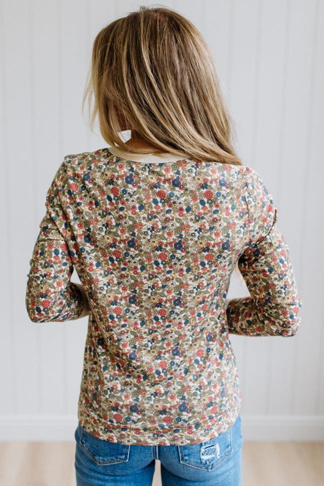 A woman viewed from behind wearing a long-sleeve floral print top with blue jeans, standing against a white paneled wall.