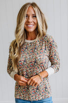 A smiling blonde woman wearing a long-sleeve crewneck top with a dense multicolor floral print on a beige background, paired with blue jeans, set against a white paneled wall.