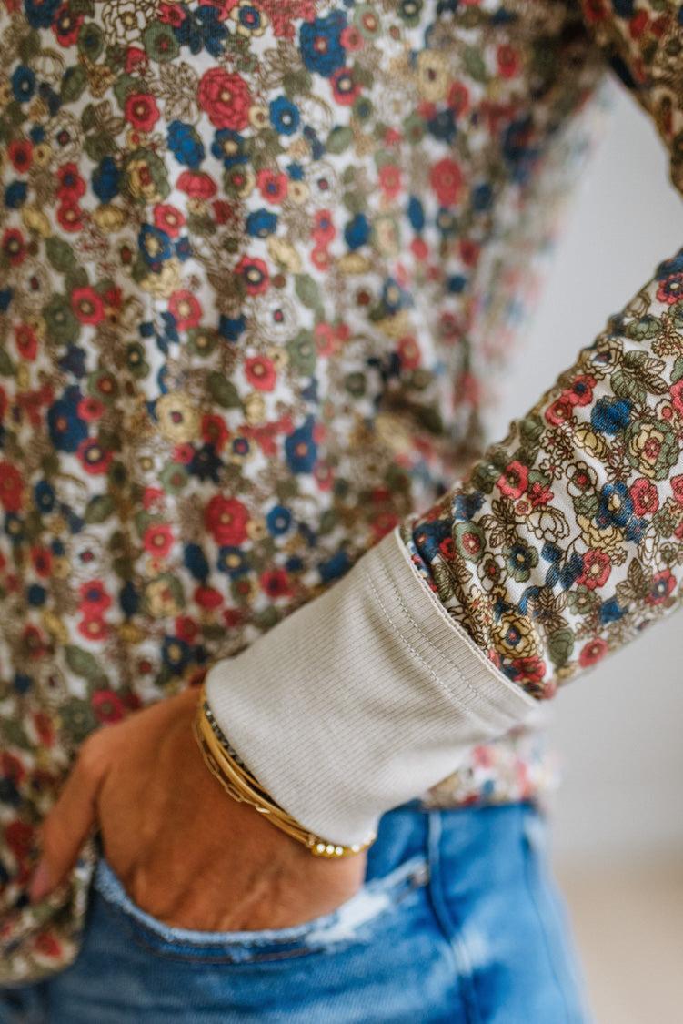 Close up shot of the sleeve cuff on a woman wearing a long-sleeve crewneck top with a dense multicolor floral print on a beige background, paired with blue jeans, set against a white paneled wall.
