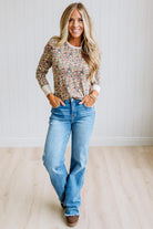 A woman stands in a bright room wearing a long-sleeve crewneck top with a dense multicolor floral print over a light beige base, paired with light-wash blue jeans and brown boots.