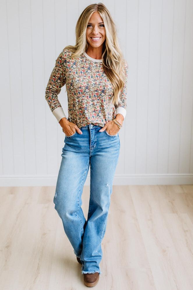 A woman stands in a bright room wearing a long-sleeve crewneck top with a dense multicolor floral print over a light beige base, paired with light-wash blue jeans and brown boots.