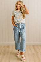 Woman wearing a floral top and light blue jeans standing against a white paneled wall.