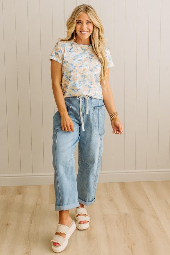 Woman wearing a floral top and blue jeans standing against a beige wall.