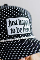 BLONDE WOMAN WEARING A BLACK AND WHITE POLKA DOT HAT WITH WHITE ROPE ACROSS THE FRONT AND A PATCH THAT SAYS JUST HAPPY TO BE HERE ON THE CENTER FRONT.