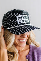 BLONDE WOMAN WEARING A BLACK AND WHITE POLKA DOT HAT WITH WHITE ROPE ACROSS THE FRONT AND A PATCH THAT SAYS JUST HAPPY TO BE HERE ON THE CENTER FRONT.