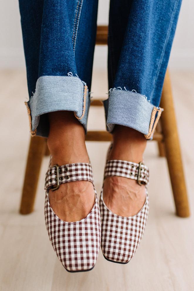 Square toed flat mary janes on a womans feet, sitting on a wooden stood with dark cuffed jeans.