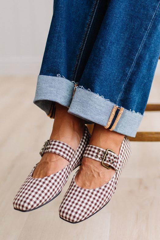 Square toed flat mary janes on a womans feet, sitting on a wooden stood with dark cuffed jeans.