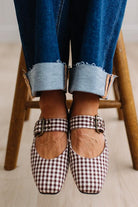 Square toed flat mary janes on a womans feet, sitting on a wooden stood with dark cuffed jeans.