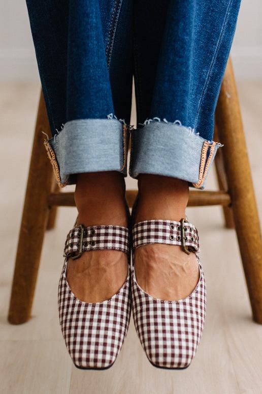 Square toed flat mary janes on a womans feet, sitting on a wooden stood with dark cuffed jeans.