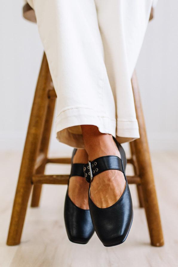 A person wearing black leather Mary Jane flats with silver-tone buckle straps, seated on a wooden stool with white pants visible against a bright indoor background.
