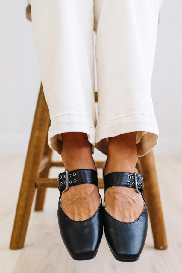 Black leather Mary Jane flats with silver buckle details, worn with off-white cuffed pants while seated on a wooden stool.