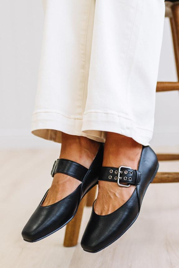 Black leather Mary Jane flats with a buckle strap, shown on a person's feet wearing off-white pants, sitting on a wooden chair.