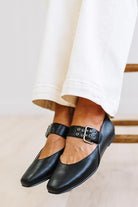A close-up of a person's feet wearing black leather Mary Jane flats with silver-tone buckle straps, paired with white pants, sitting on a wooden chair.
