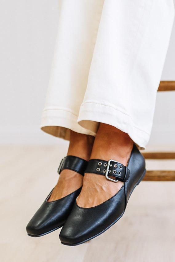 A close-up of a person's feet wearing black leather Mary Jane flats with silver-tone buckle straps, paired with white pants, sitting on a wooden chair.