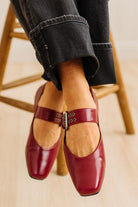 Red loafers with a wooden sole worn by a person sitting on a wooden stool.
