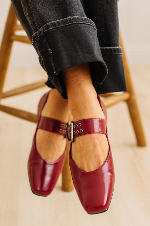 Red loafers with a wooden sole worn by a person sitting on a wooden stool.