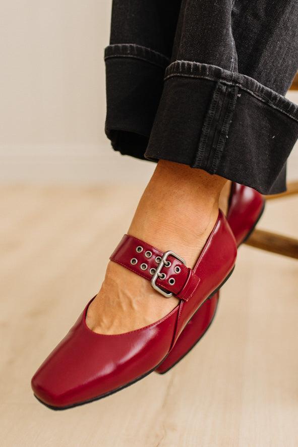 Red loafers with a silver buckle worn with black jeans on a beige surface.