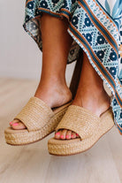 Woven sandals worn with a patterned dress on a light wooden floor.