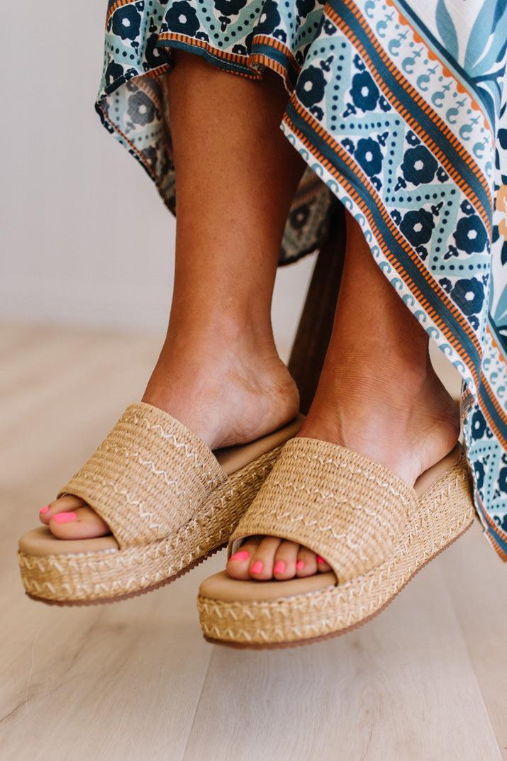 Woven sandals worn with a patterned dress on a light wooden floor.