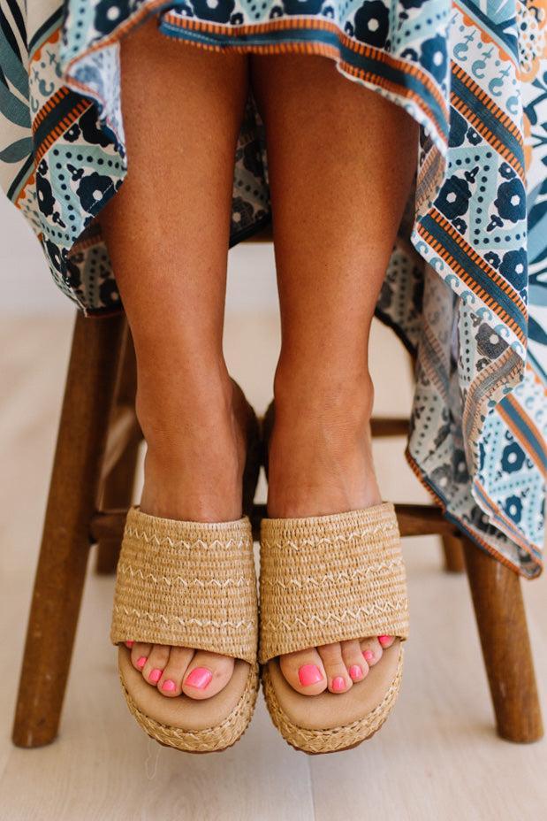 Person wearing beige sandals with a patterned dress and chair in the background