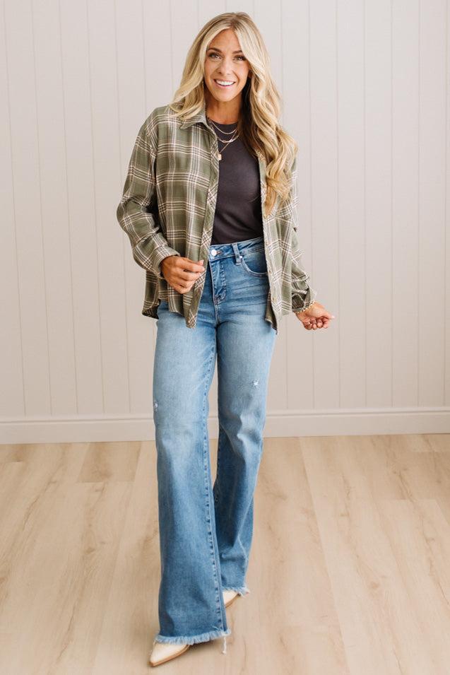 A woman modeling a green and brown plaid flannel shirt over a dark gray top and light blue flare jeans, standing in a room with light wood floors and white paneled walls.