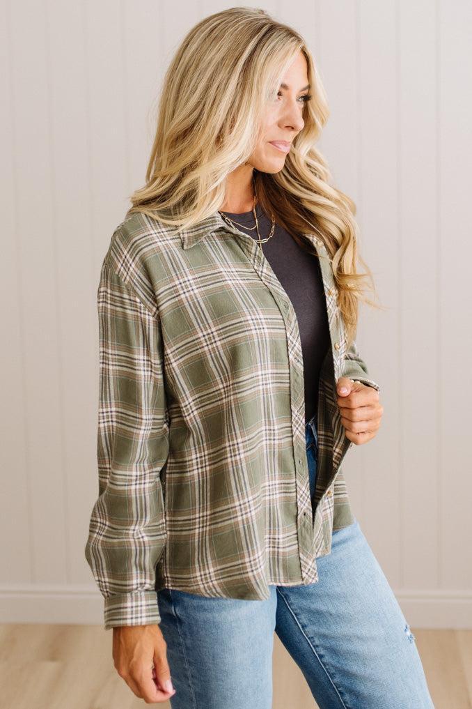 A woman wearing a sage green plaid button-up shirt over a black top and blue jeans, standing against a light beige paneled wall.