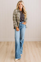 A woman modeling a pair of high-waisted, wide-leg blue jeans with a dark gray top and an olive green plaid flannel shirt, standing against a white paneled wall and light wood floor.