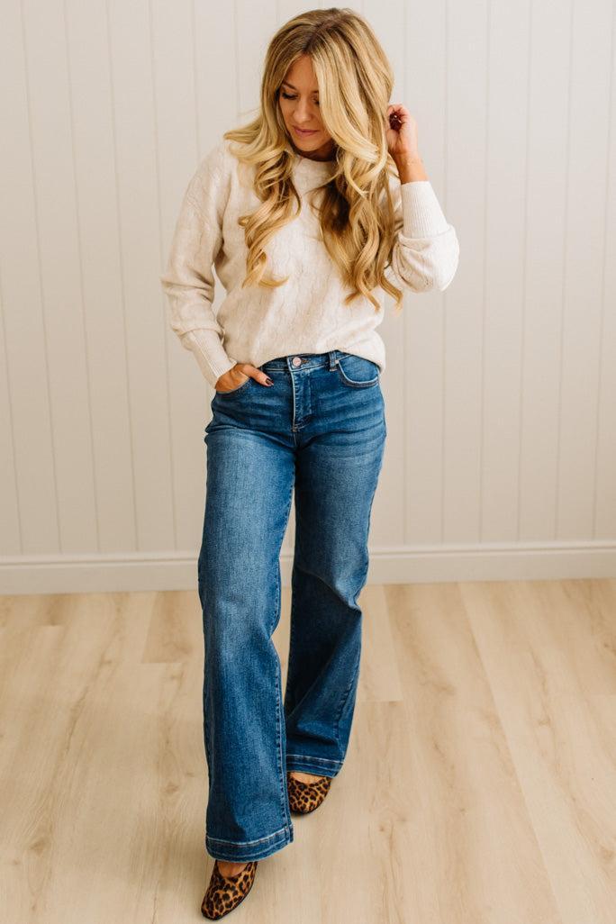 Woman wearing a white sweater and blue jeans standing against a light wooden paneled wall.