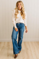 Woman wearing a beige sweater and blue jeans standing against a white paneled wall.