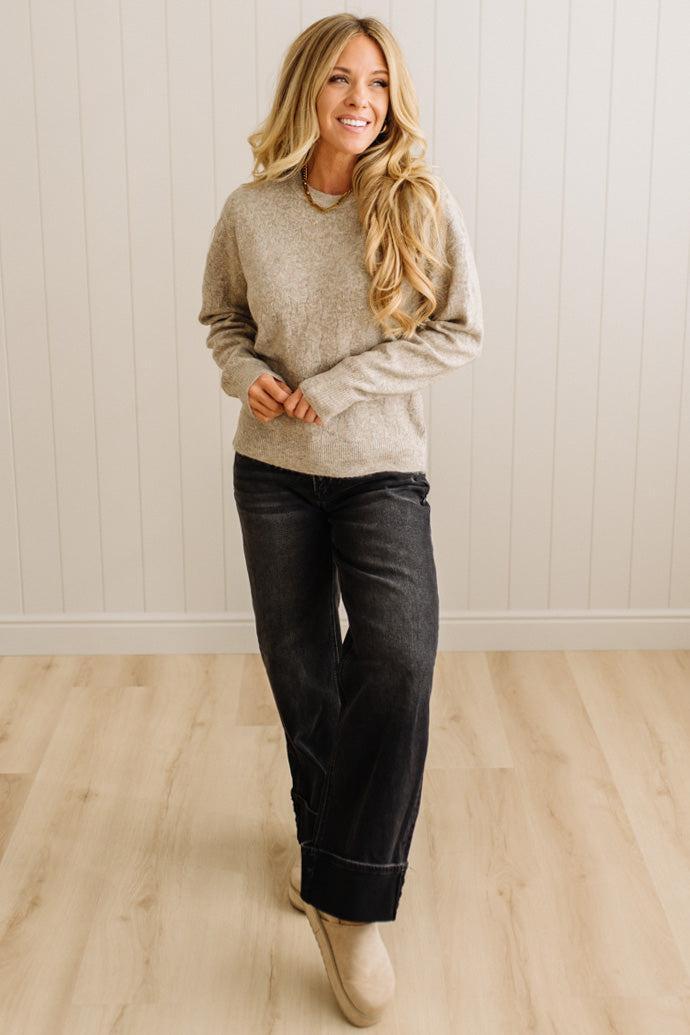 Woman wearing a beige sweater and black pants standing against a light wooden paneled wall.