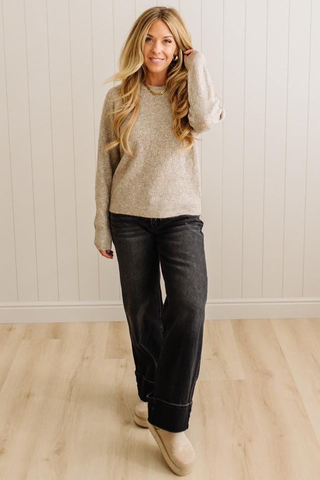 Woman wearing a beige sweater and dark jeans standing against a white paneled wall.